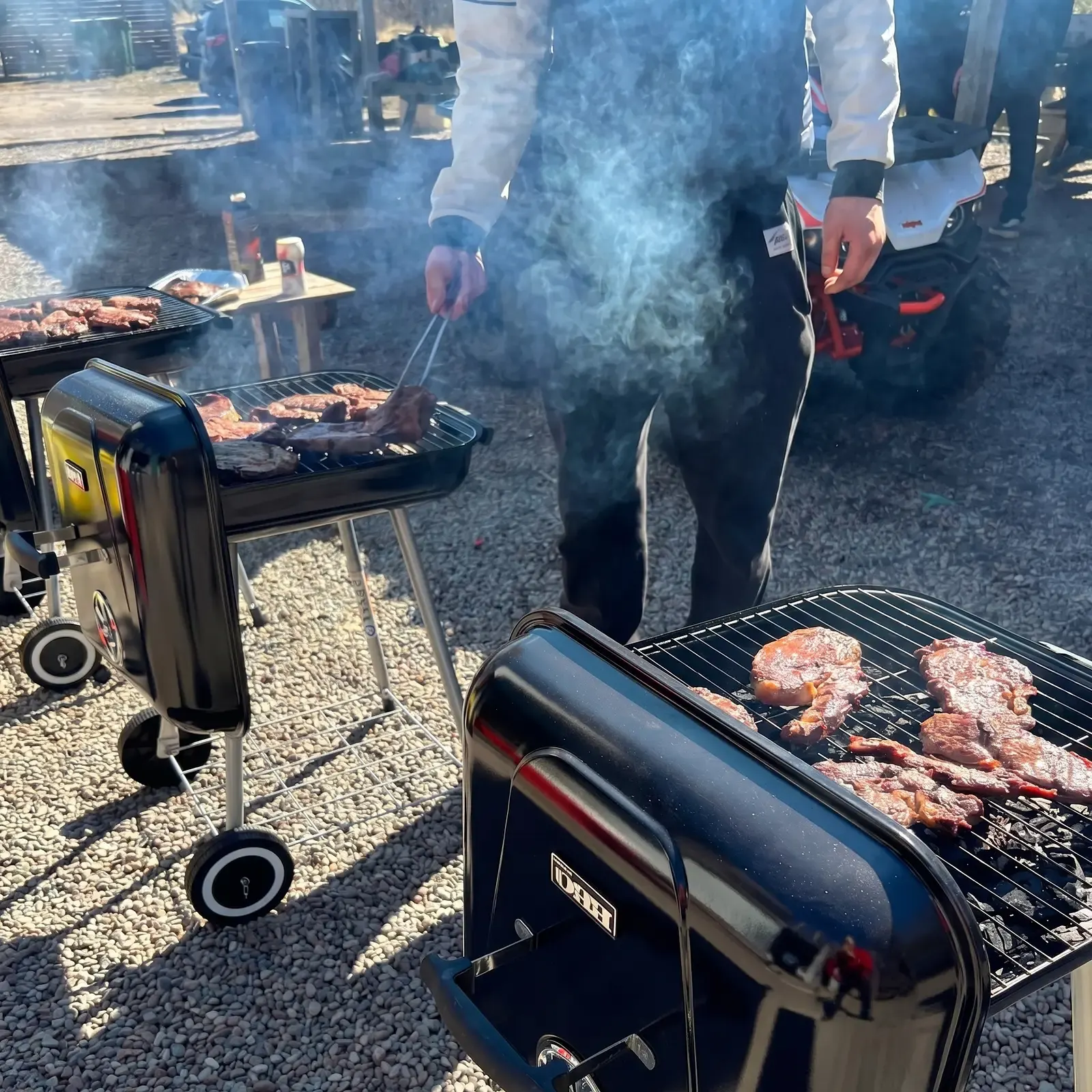Personer grillar kött på flera grillmaskiner utomhus med rök.