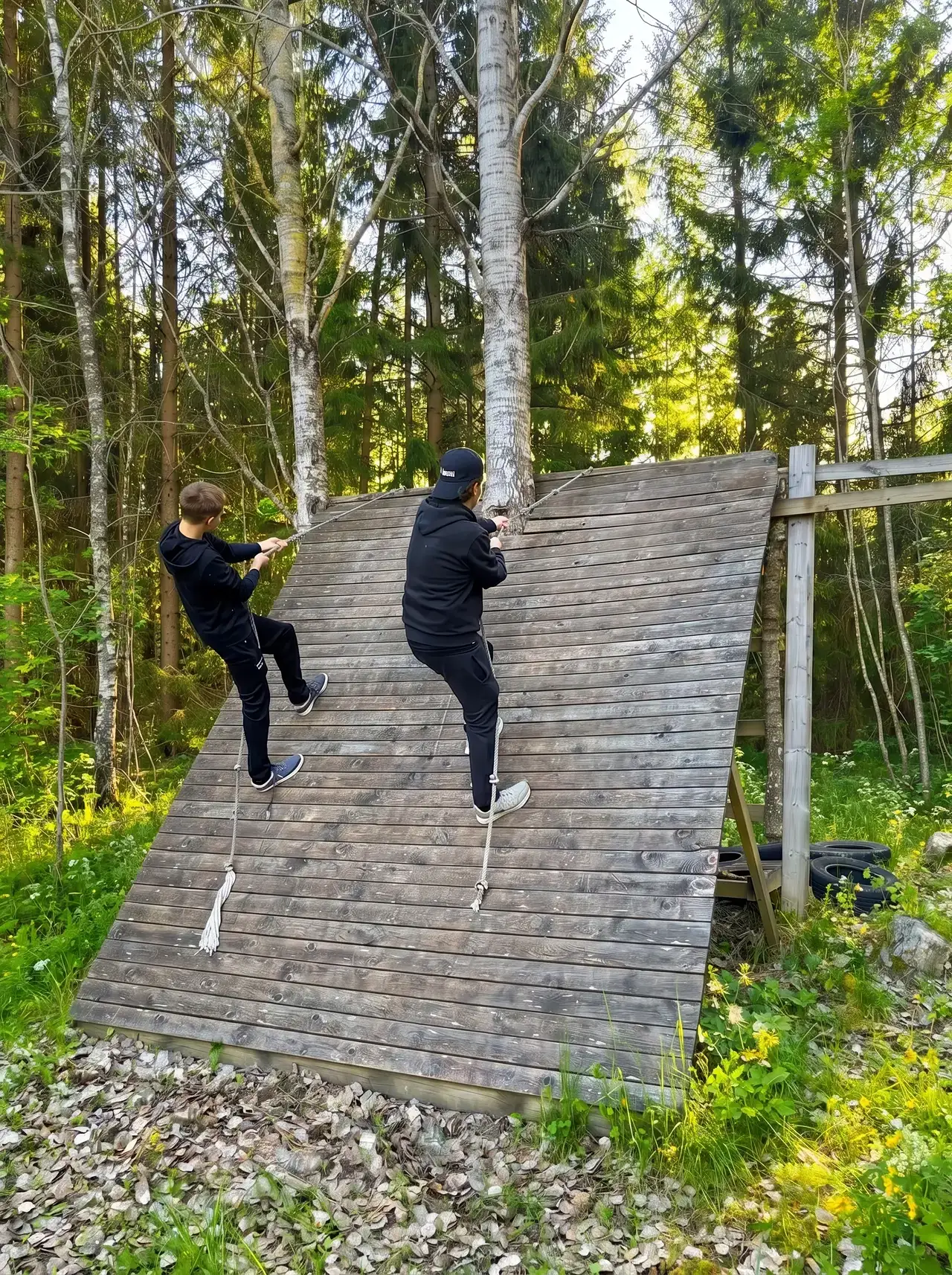 Två personer klättrar på en lutande trävägg i skogen.